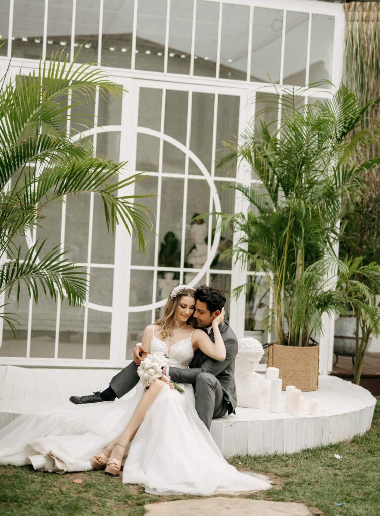 Brudpar sitter tätt tillsammans framför glasdörrar och palmer